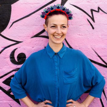 A white woman standing with her hands on her hips staring beyond the camera. She wears a colourful halo style, graphic stripe festival headdress and a blue shirt in front of a wall covered in pink graffiti