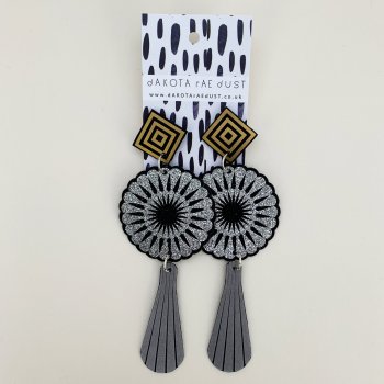 A pair of extra long art deco earrings featuring black and silver circular motifs and elongated grey teardrops suspended below black and gold square studs are seen against an off white background