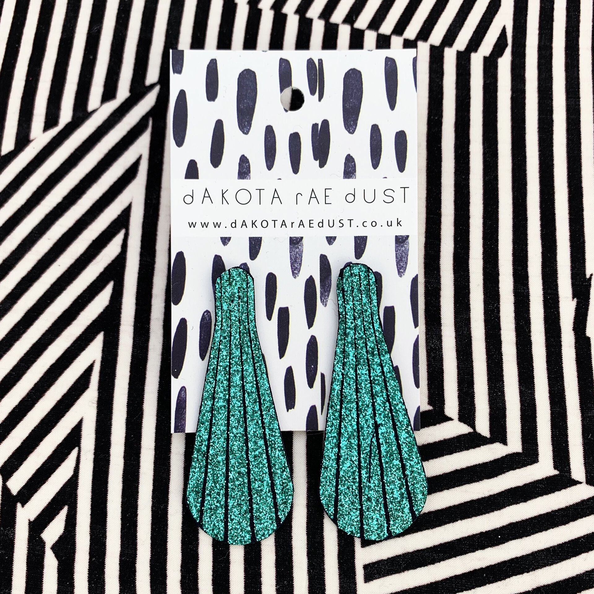 A pair of elongated emerald green glittery studs mounted on a black and white patterned, dakota rae dust branded card are sitting on a boldly graphic striped black and white background