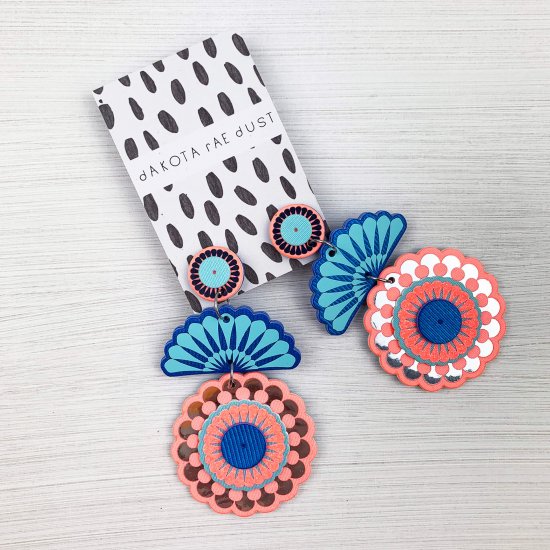 A pair of Statement stud circle arch earrings cut from peach and turquoise fabric and printed with silver detail are displayed on a black and white patterned dakota rae dust branded card are lying on an off white background.