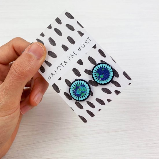 A pair of small circle studs in navy and light blue are mounted on a black and white patterned, dakota rae dust branded card and held between the thumb and fore finger of a woman's hand