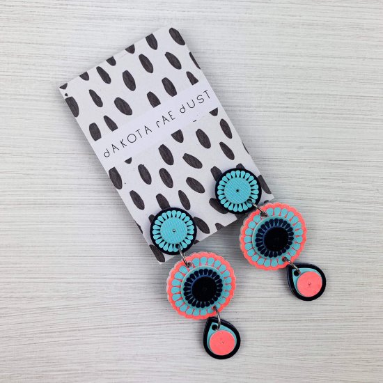 Dangly charm earrings in coral and blue displayed on a monochrome patterned backing card