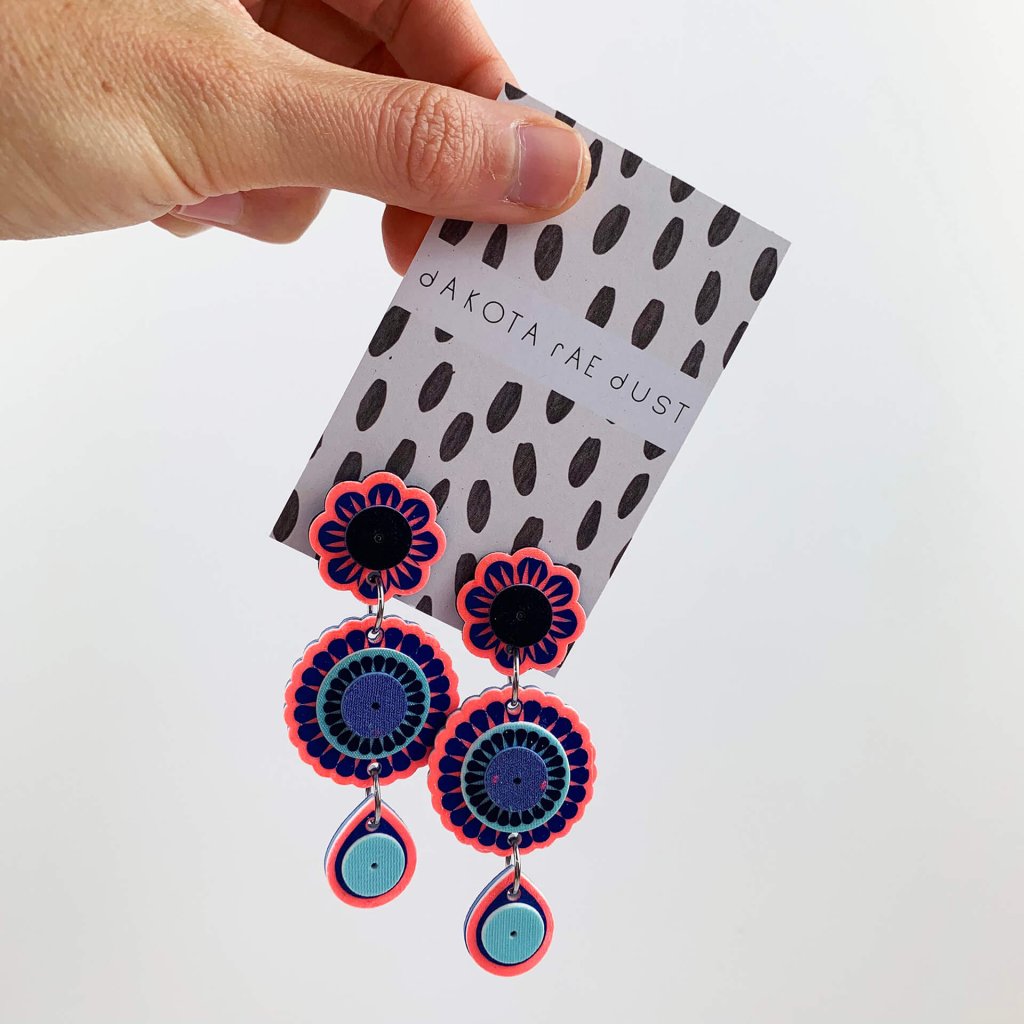 A pair of neon coral and blue dangly charm earrings mounted on a black and white patterned, dakota rae dust branded card are held at a jaunty angle by a hand, just visible in the top left hand corner on the image