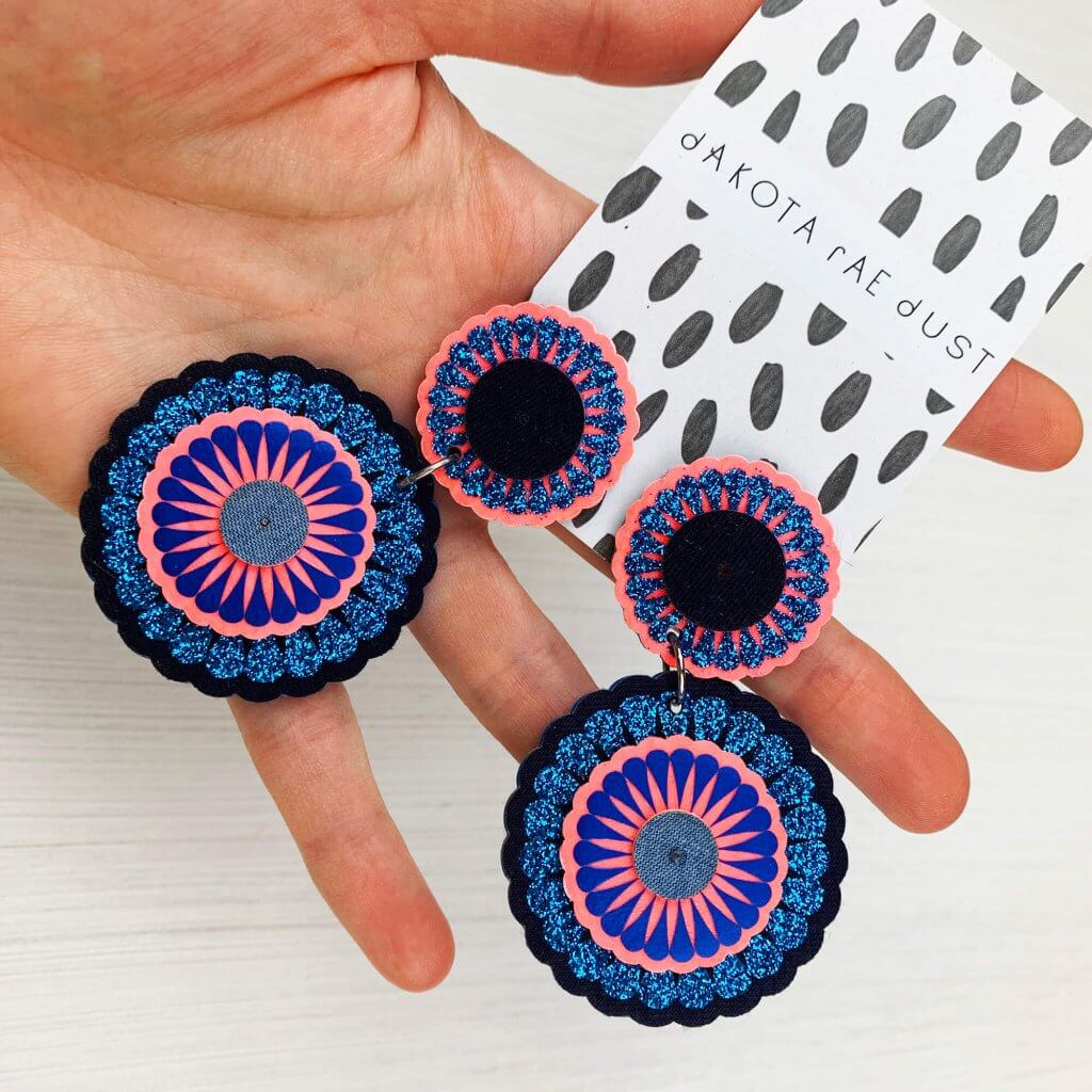Glittery blue and pink decorative disc statement earrings held in a woman's hand against a white background