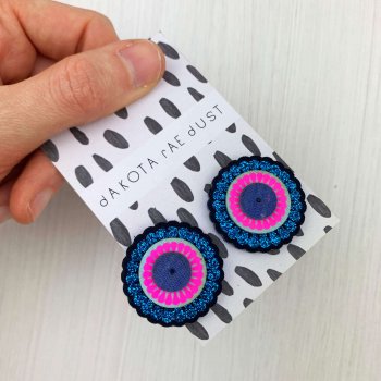 Oversize stud earrings in bright blue glittery and fluorescent pink, on a black and white patterned backing card, held in a hand over a textured white backdrop.