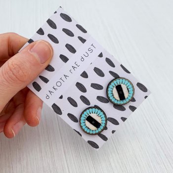 A pair of circular, gold and blue Small stripe stud earrings on a monochrome patterned backing card held against a clean white background by the thumb and forefingers of a woman's hand..