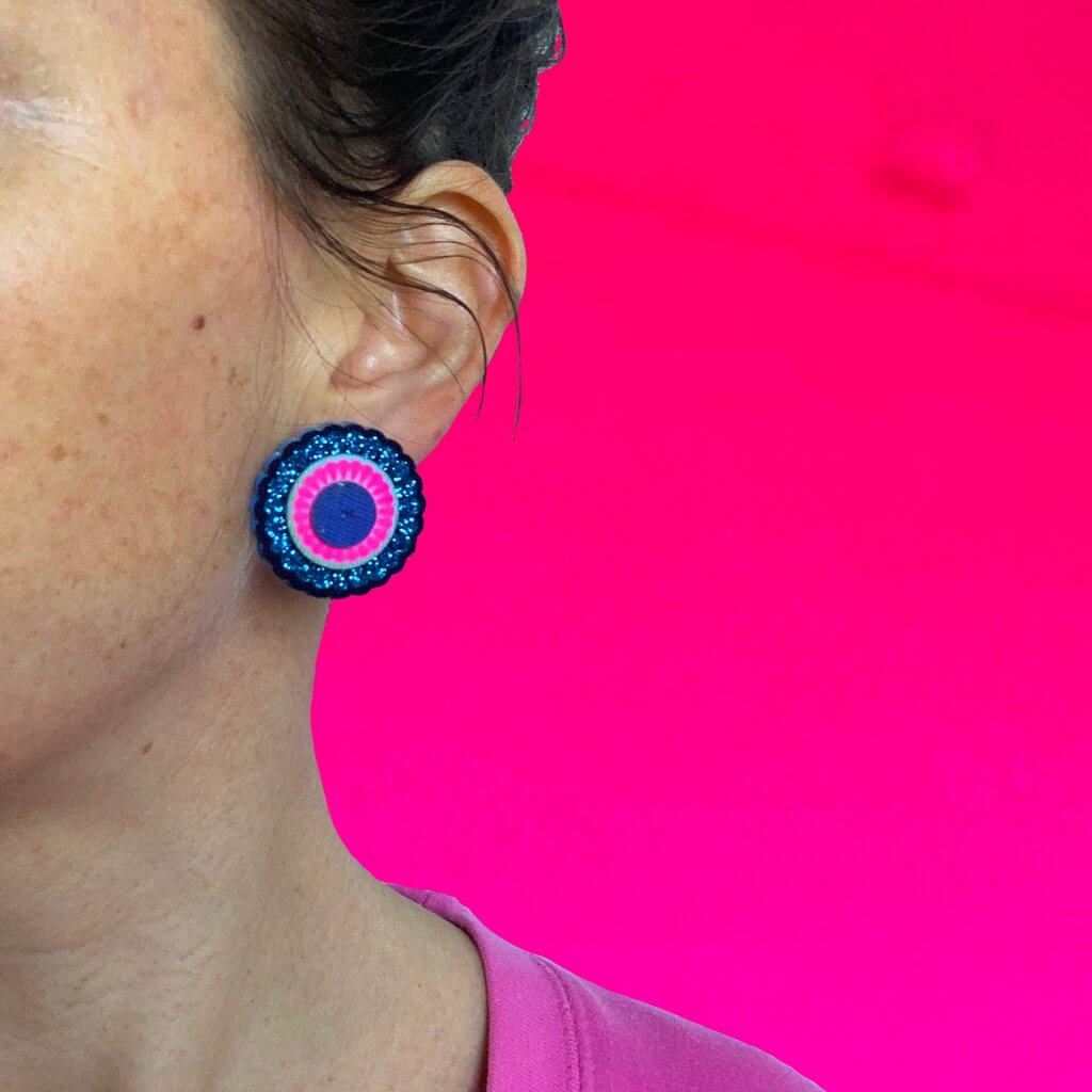 A close up on a circular gittery blue and neon pink stud earring, worn by a woman in a pink sweatshirt against a bright pink backdrop.