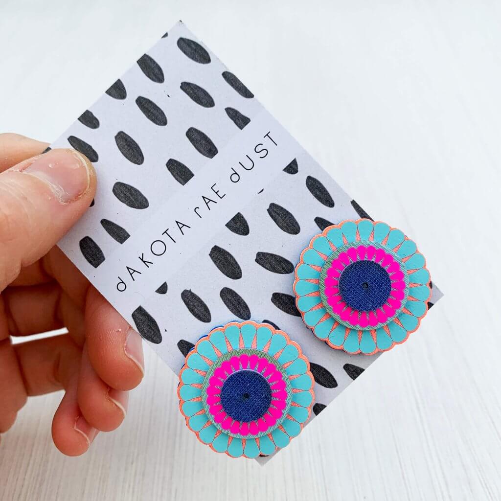 A colourful pair of circular, ornate oversize studs in blue, peach and neon pink, made of tiers of laser cut and vinyl printed discs are seen mounted on a monochrome patterned dakota rae dust branded card, held in a woman's hand. only the fingers and thumb are visible.