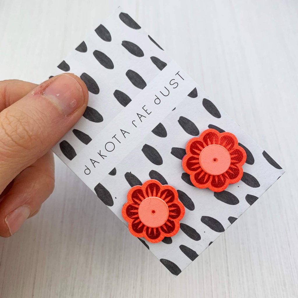 A pair of red, orange and peach flower stud earrings mountede on a black and white patterned, dakota rae dust branded card between a just visible thumb and forefinger