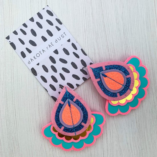 A pair of decorative teardrop shaped, oversize earrings in pink, turquoise and gold mounted on a black and white patterned dakota rae dust branded card, photographed against an off white background