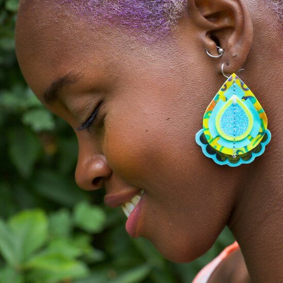 A close up of a young black woman's profile. Her head is lowered and she is smiling. A large teardrop shaped earring in brirght yellow and blue is the main focus of the photo.