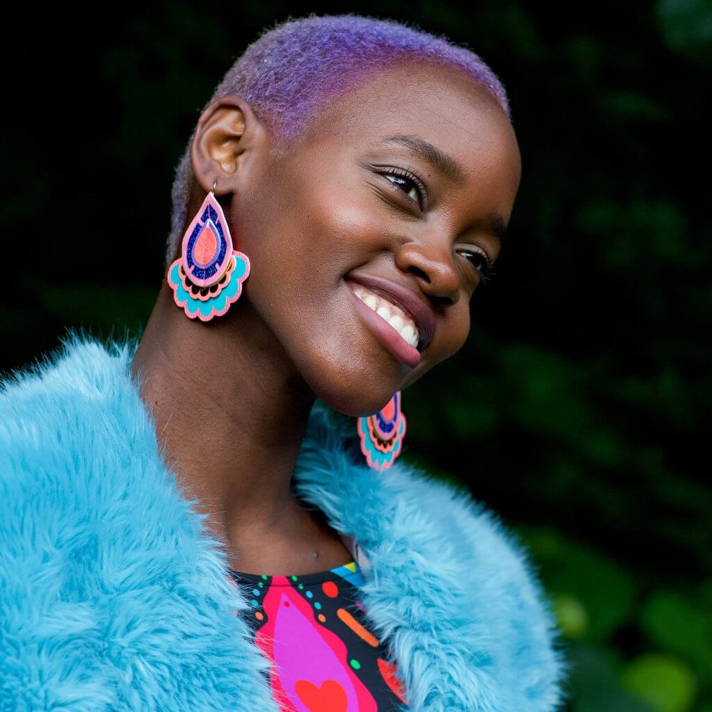A young black woman with cropped lilac hair wears a pair of colourful, oversize statement earrings by dakota rae dust and a bright blue faux fur wrap. She is smiling and looking past the camera.