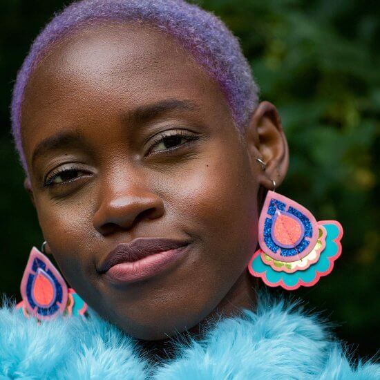 A young black woman with short lilac hair wearing a blue furry collar and colourful statement earrings smiles and looks into the camera