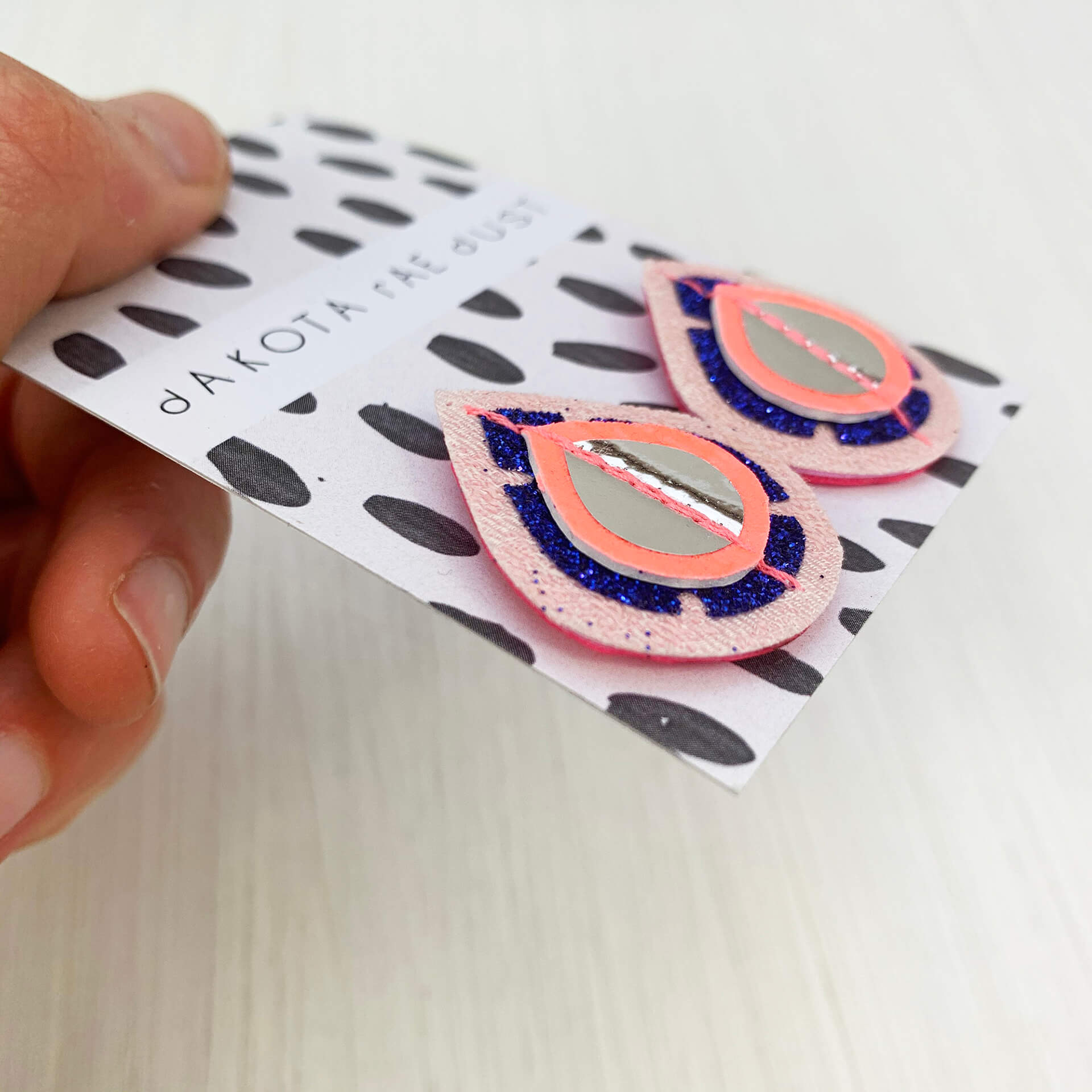 A pair of recycled pink kimono fabric and glittery blue vinyl teardrop shaped studs mounted on a dakota rae dust branded card are held between a just visible thumb and forefinger.