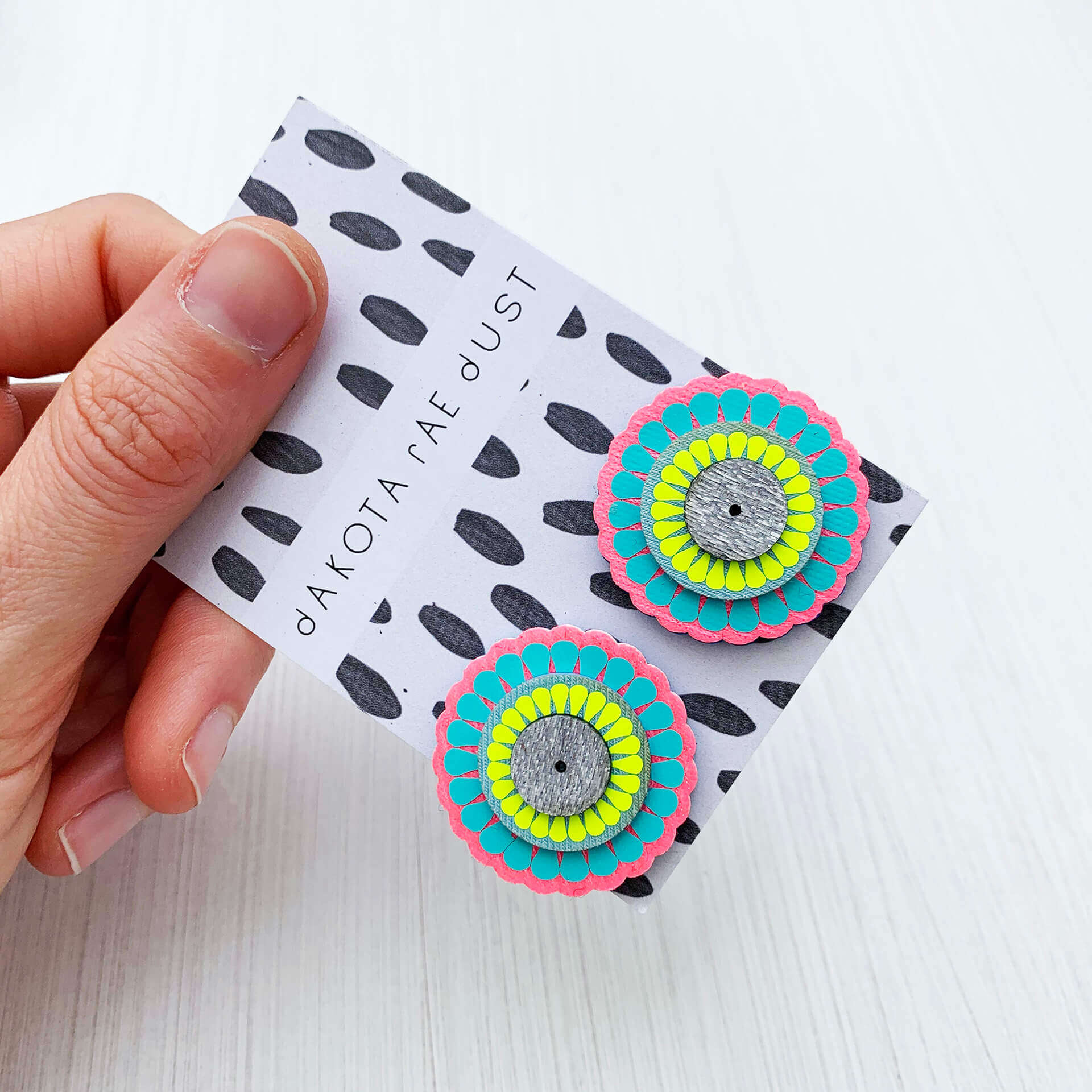 A pair of colourful ornately patterned, oversize studs, backed on a black and white patterned swing tag, printed with a dakota rae dust logo are held in a woman's hand. Only the thumb and fingers are visible. The background is plain off white. The studs are circle shapes in turquoise, pink and yellow.