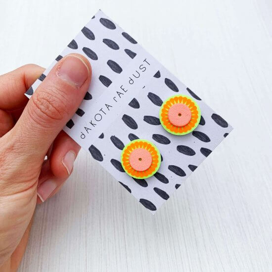 A pair of small stud earrings in zingy fluorescent yellow and orange, backed on a black and white patterned, dakota rae dust branded card are held by a woman's hand against an off white background.