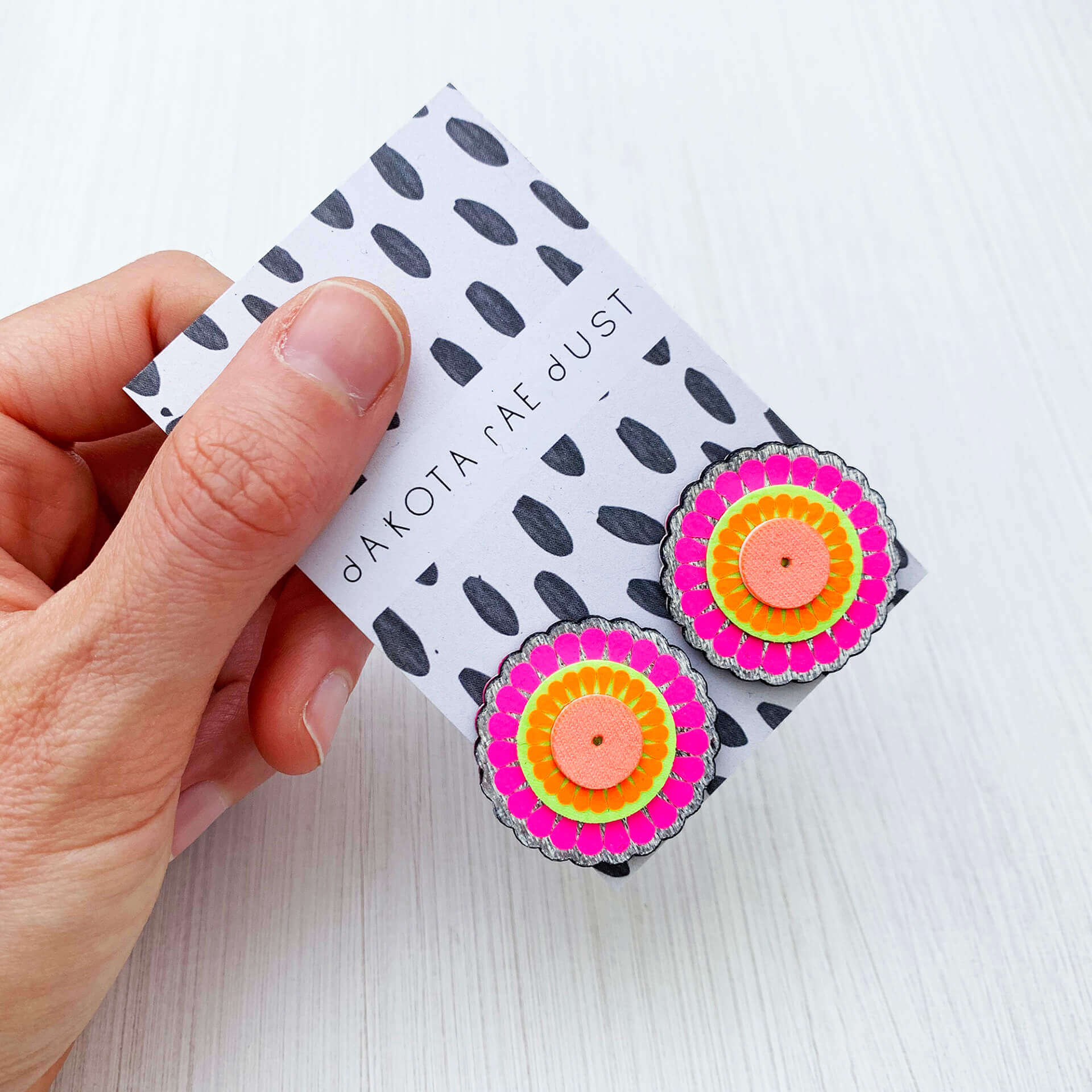 Bright pink and orange, colourful oversize studs, mounted on a black and white patterned swing tag, printed with the dakota rae dust logo. The earrings are held in a hand over an off white background. only the thumb and fingers are visible.