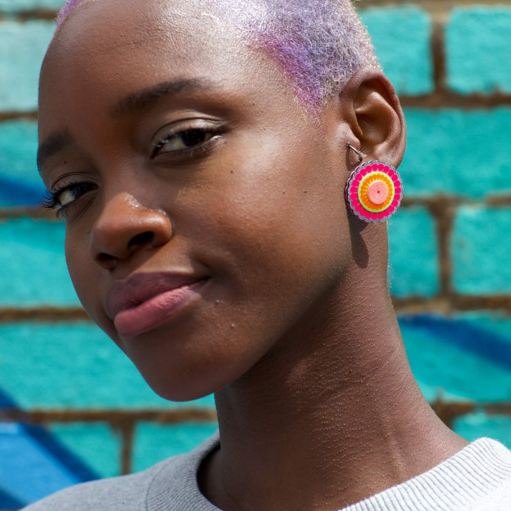 A young black woman with short lilac hair wears a pair of neon pink, circular oversize stud earrings against a bright turquoise wall.