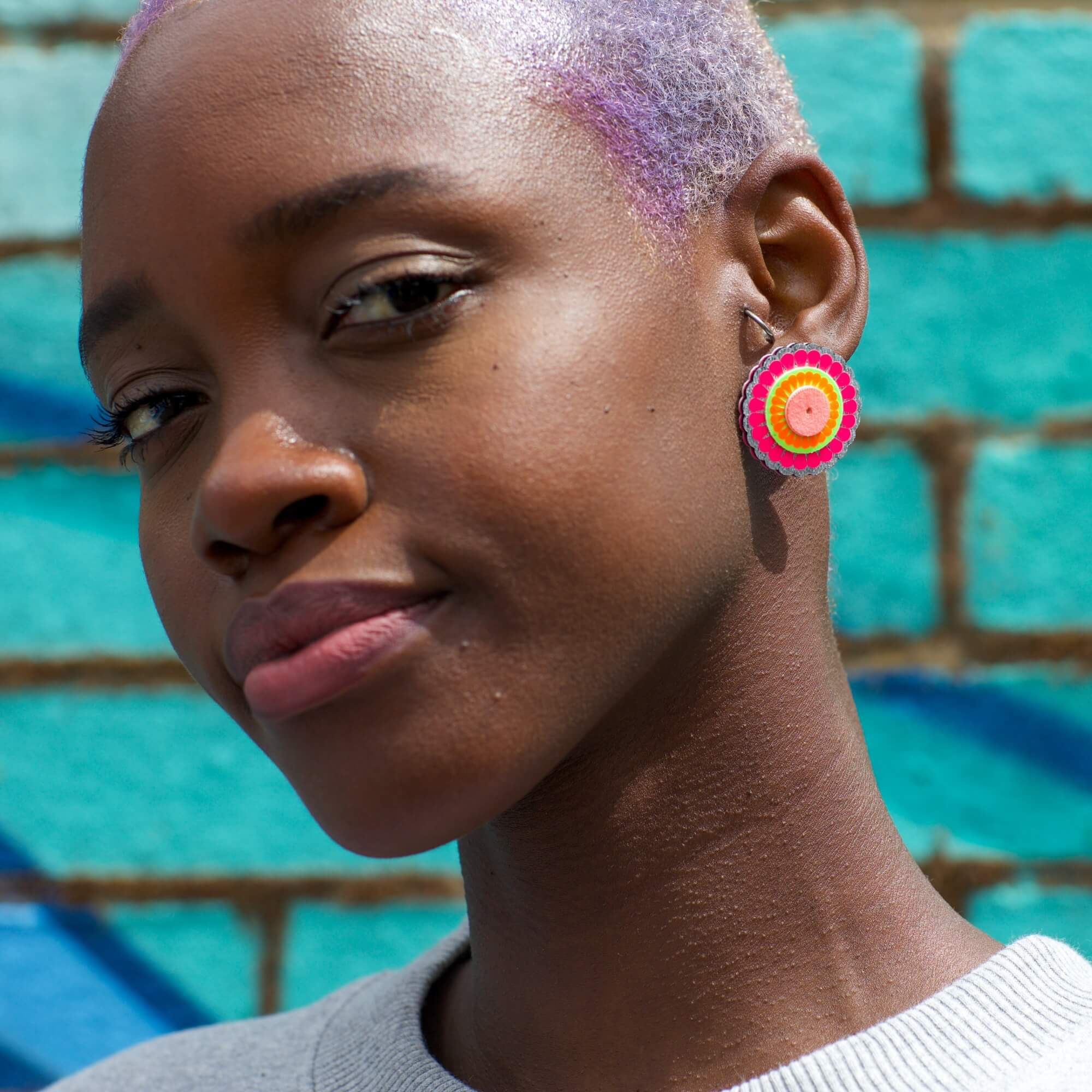 A young black woman with very short lilac hair is gazing into the camera. she is wearing a pair of large circular stud earrings and standing against a bright turquoise brick wall