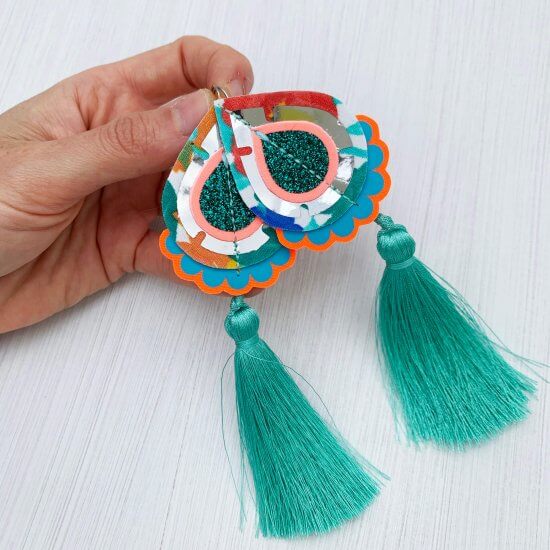 a pair of teardrop shape statement earrings with silky turquoise tassels are held between a woman's thumb and fore finger