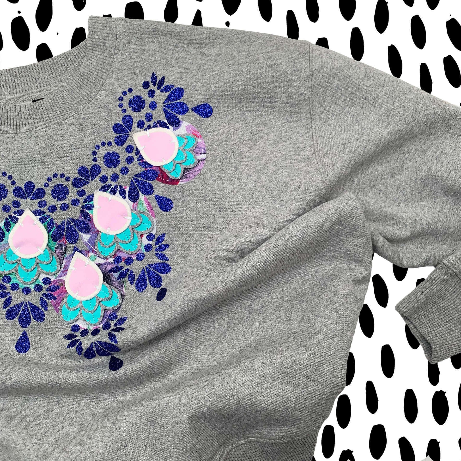 An embellished sweatshirt in grey, glittery blue, turquoise and pale pink laid out against a black and white patterned background. Only the right hand side of the garment is in shot. The grarment is a grey marl dropped shoulder sweatshirt.