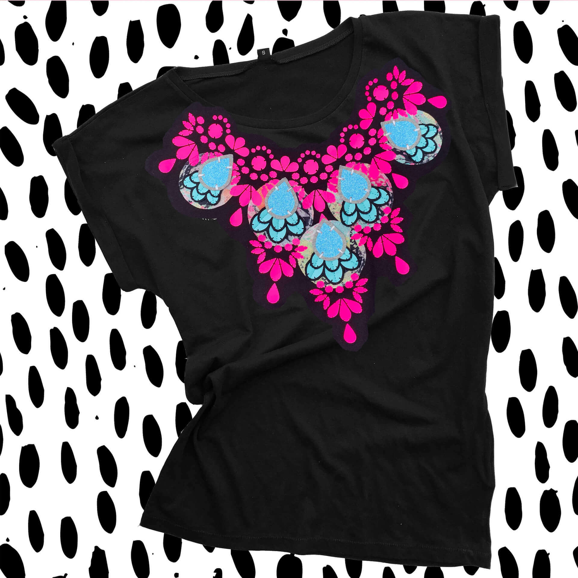 An embellished slim fit T-shirt in black, fluorescent pink and blue, laid flat against a black and white patterned background.