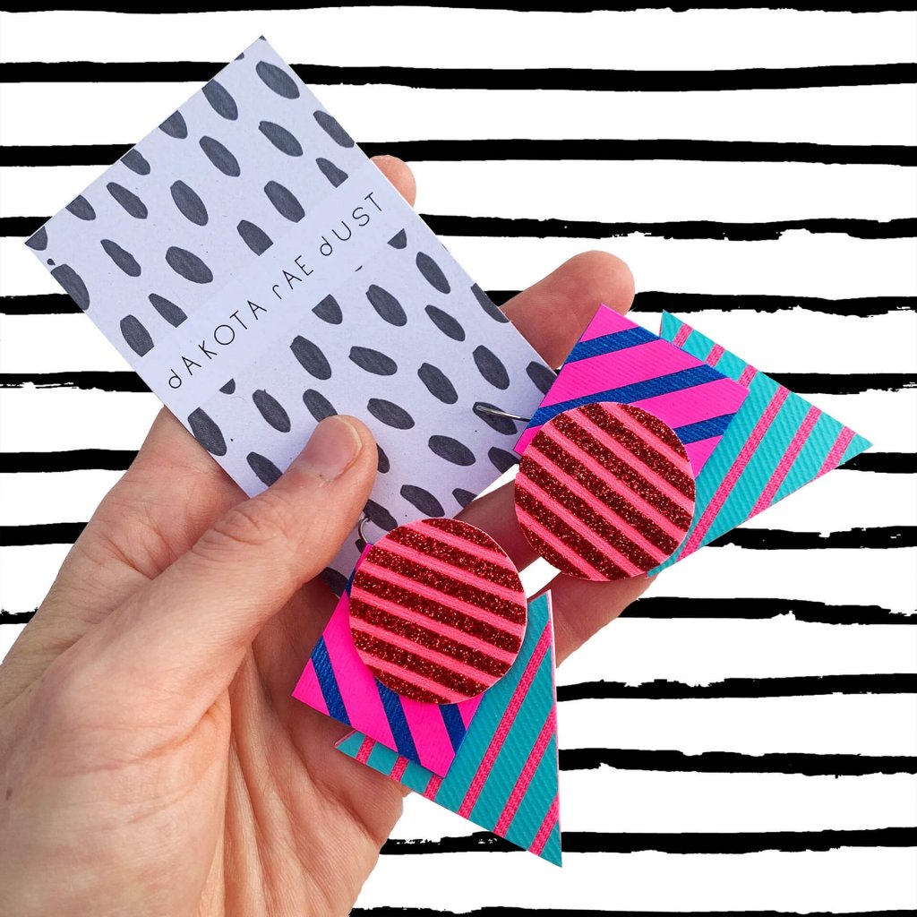 A pair of colourful dakota rae dust Triangular stripe earrings, featuring a cluster of three geometric shapes printed with bold graphic stripes displayed on a black and white patterned, dakota rae dust branded card, held in a white woman's hand with a black and white striped background