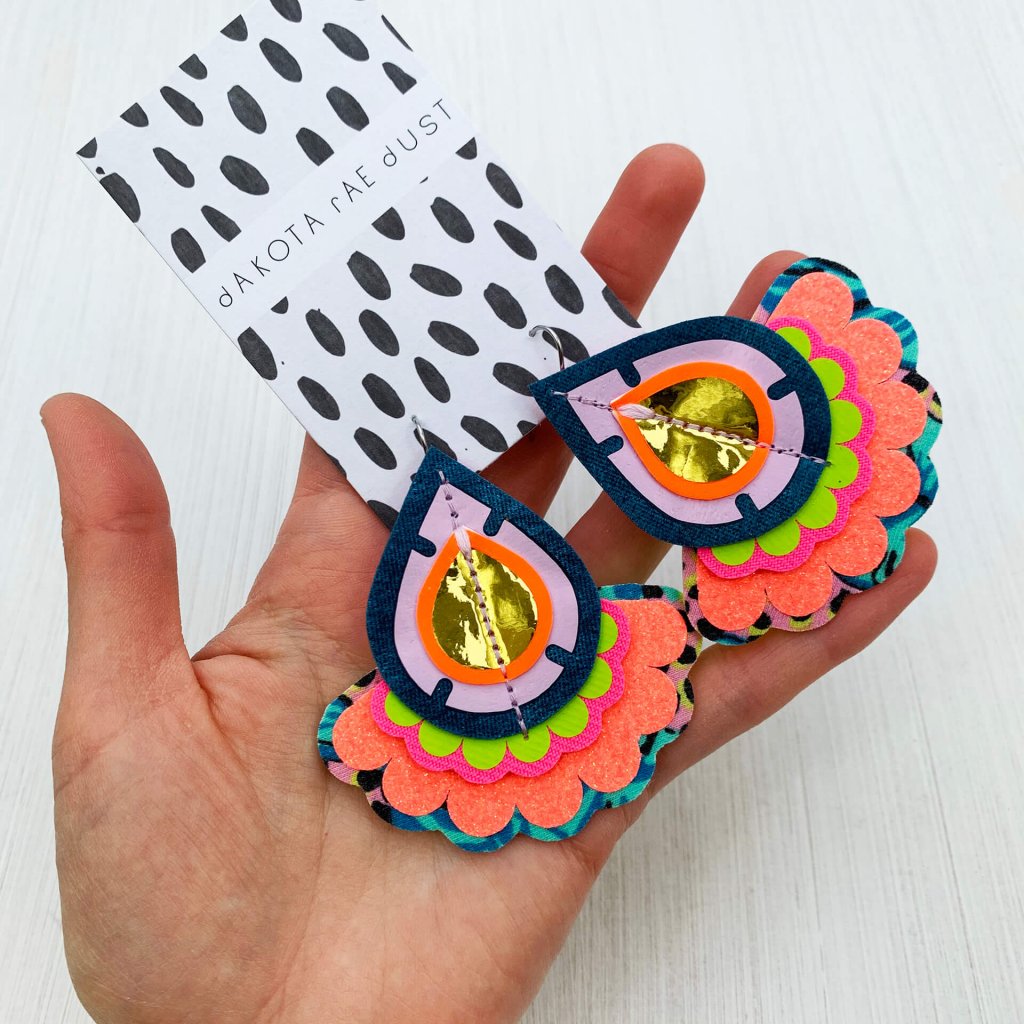 A pair of colourful oversize teardrop earrings mounted on a dakota rae dust branded card in a white woman's hand