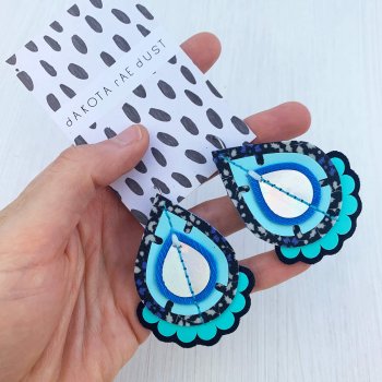 A pair of decorative teardrop earrings in shades of blue and silver mounted on a black and white patterned dakota rae dust branded card, held in a woman's hand against an off white background