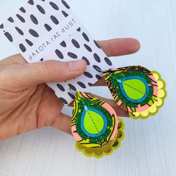 A pair of green, lime, peach and gold patterned fabric earrings, mounted on a black and white patterned dakota rae dust branded card, against an off white background
