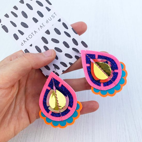 A pair of glittery teardrop earrings in pink, blue and gold, mounted on a black and white patterned dakota rae dust branded card are held in a woman's hand against an off white background