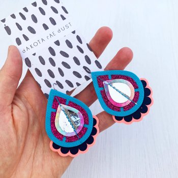 A pair of turquoise and pink colourful statement earrings, mounted on a black and white patterned dakota rae dust branded card are held in a woman's hand against an off white background