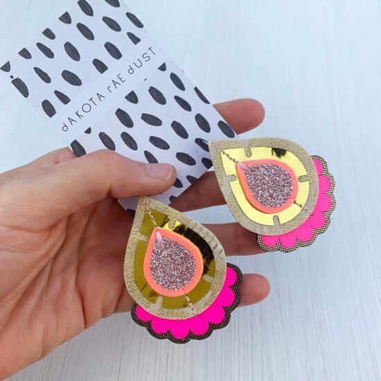 A pair of shiny gold and fluorescent pink teardrop shaped earrings, backed on a black and white patterned branded card are held in a woman's hand against an off white background