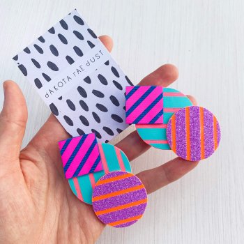 A pair of colourful stripey cluster earrings, featuring boldly striped geometric shapes in pink, blue, turquoise and purple glitter, displayed on a black and white patterned, dakota rae dust branded card, held in a woman's hand.