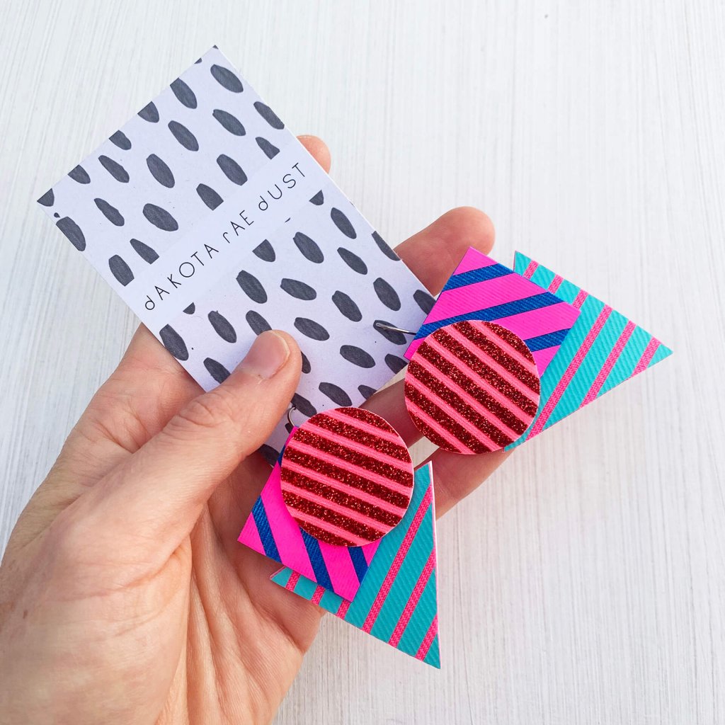 A pair of colourful Triangular stripe earrings, featuring a cluster of three geometric shapes printed with bold graphic stripes displayed on a black and white patterned, dakota rae dust branded card, held in a white woman's hand with an off white background