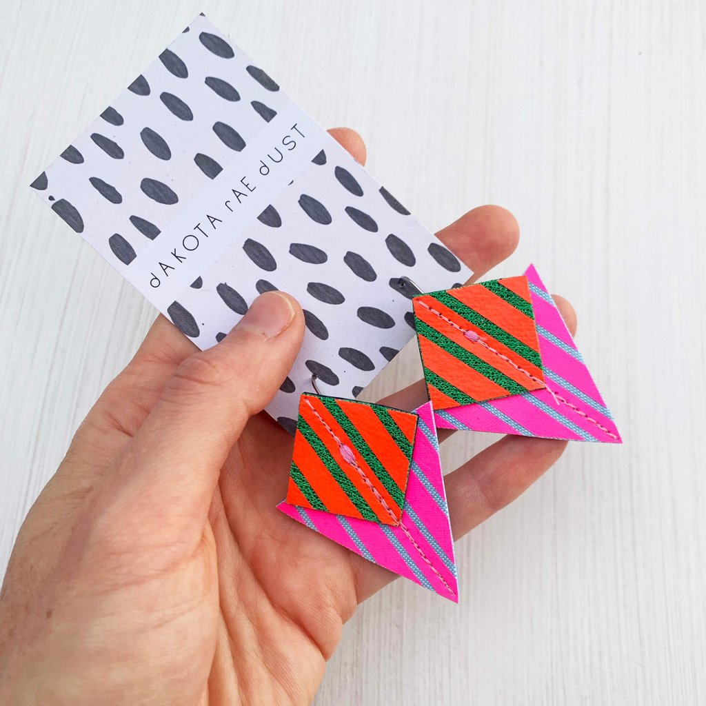 A pair of fluorescent pink, red and emerald green Stripey triangle earrings, featuring a triangle and square printed with bold graphic stripes displayed on a black and white patterned, dakota rae dust branded card, held in a white woman's hand against an off white background