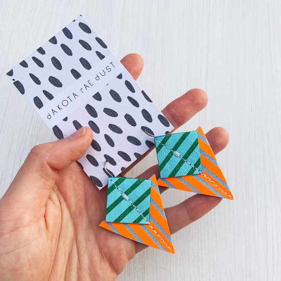 A pair of fluorescent orange, green and blue Stripey triangle earrings, featuring a triangle and square printed with bold graphic stripes displayed on a black and white patterned, dakota rae dust branded card, held in a white woman's hand against an off white background
