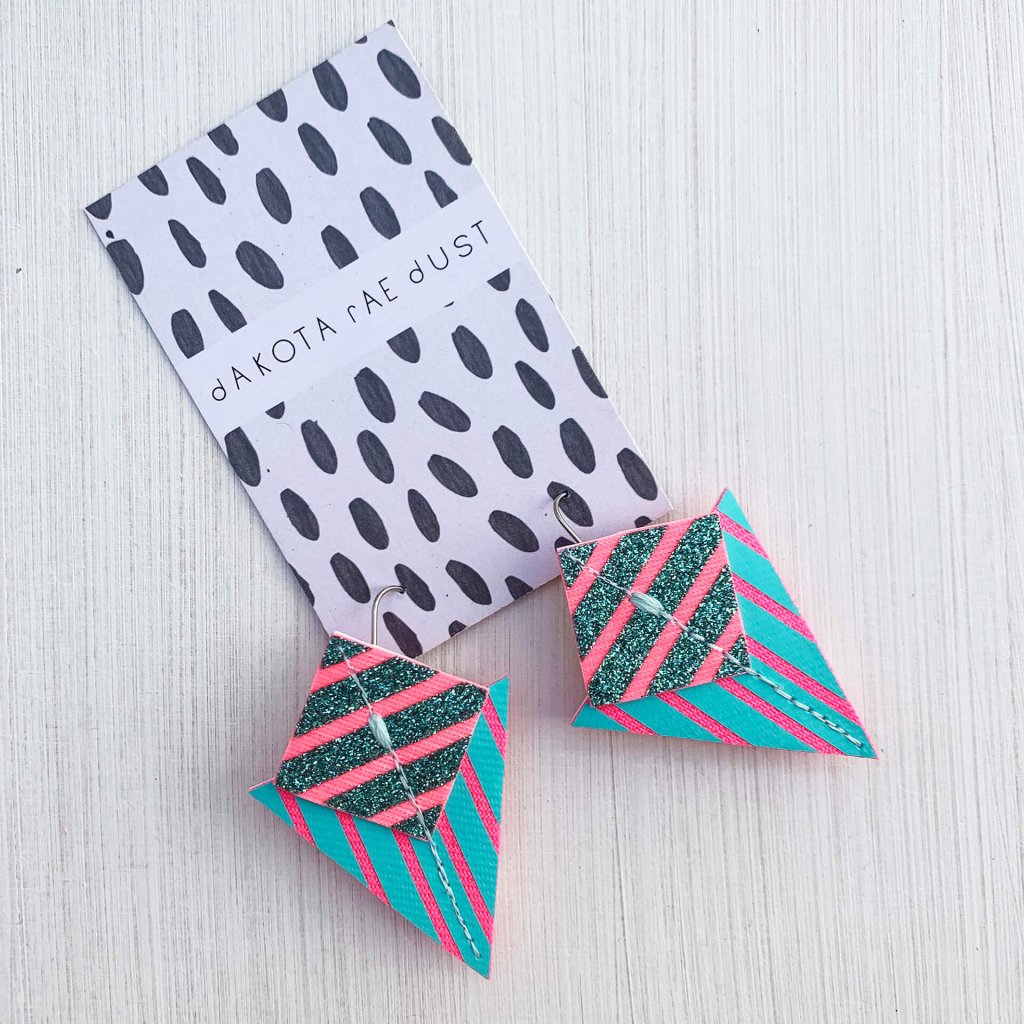 A pair of turquoise, fluorescent pink and emerald green glitter Stripey triangle earrings, featuring a triangle and square printed with bold graphic stripes displayed on a black and white patterned, dakota rae dust branded card, against an off white background