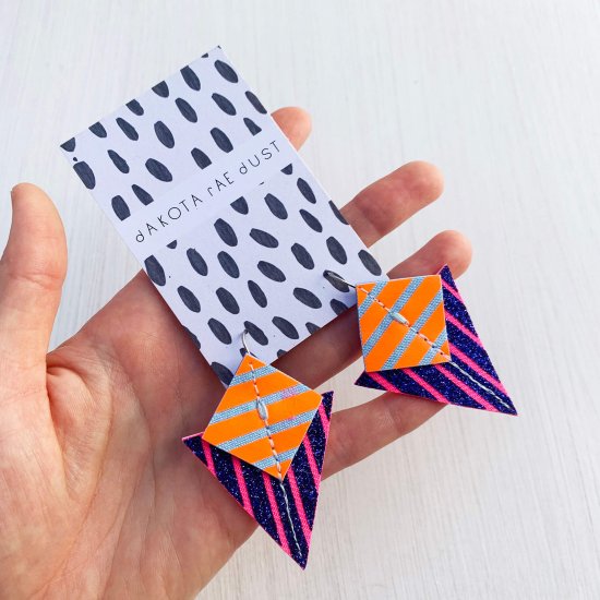 A pair of glittery blue, pink, orange and pale blue Stripey triangle earrings, featuring a triangle and square printed with bold graphic stripes displayed on a black and white patterned, dakota rae dust branded card, held in a white woman's hand against an off white background