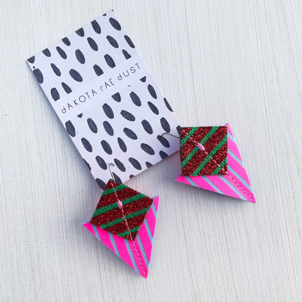 A pair of fluorescent pink, glittery red, green and pale blue Stripey triangle earrings, featuring a triangle and square printed with bold graphic stripes displayed on a black and white patterned, dakota rae dust branded card, against an off white background