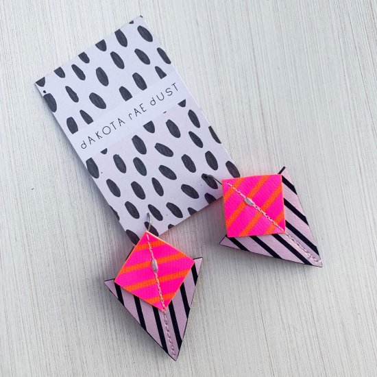 A pair of lilac, fluorescent pink, orange and navy Stripey triangle earrings, featuring a triangle and square printed with bold graphic stripes displayed on a black and white patterned, dakota rae dust branded card, against an off white background