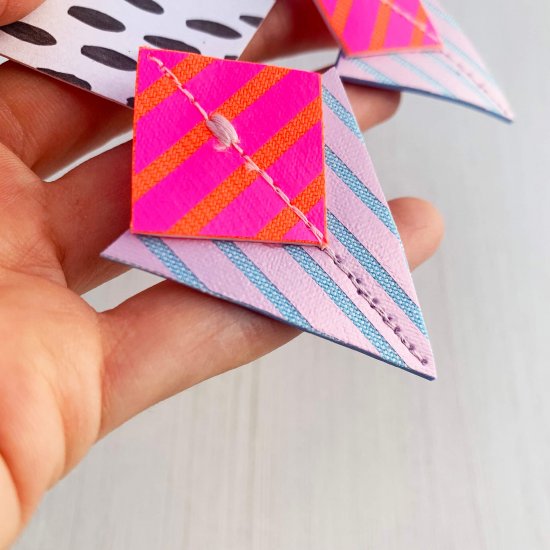 A pair of lilac, fluorescent pink, orange and pale blue Stripey triangle earrings, featuring a triangle and square printed with bold graphic stripes displayed on a black and white patterned, dakota rae dust branded card, held in a white woman's hand against an off white background