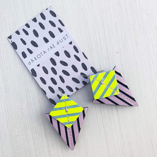 A pair of lilac, fluorescent yellow and navy Stripey triangle earrings, featuring a triangle and square printed with bold graphic stripes displayed on a black and white patterned, dakota rae dust branded card, against an off white background