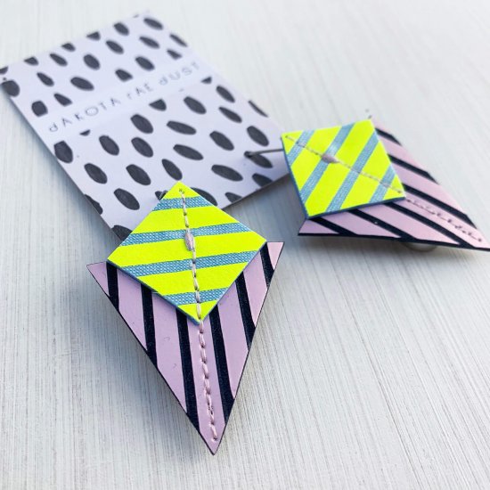 Statement stripey earrings in lilac, neon yellow and navy. A pair of earrings consisting of a stripey square and triangle component are seen mounted on a mononchrome patterned dakota are dust card against a white textured background.