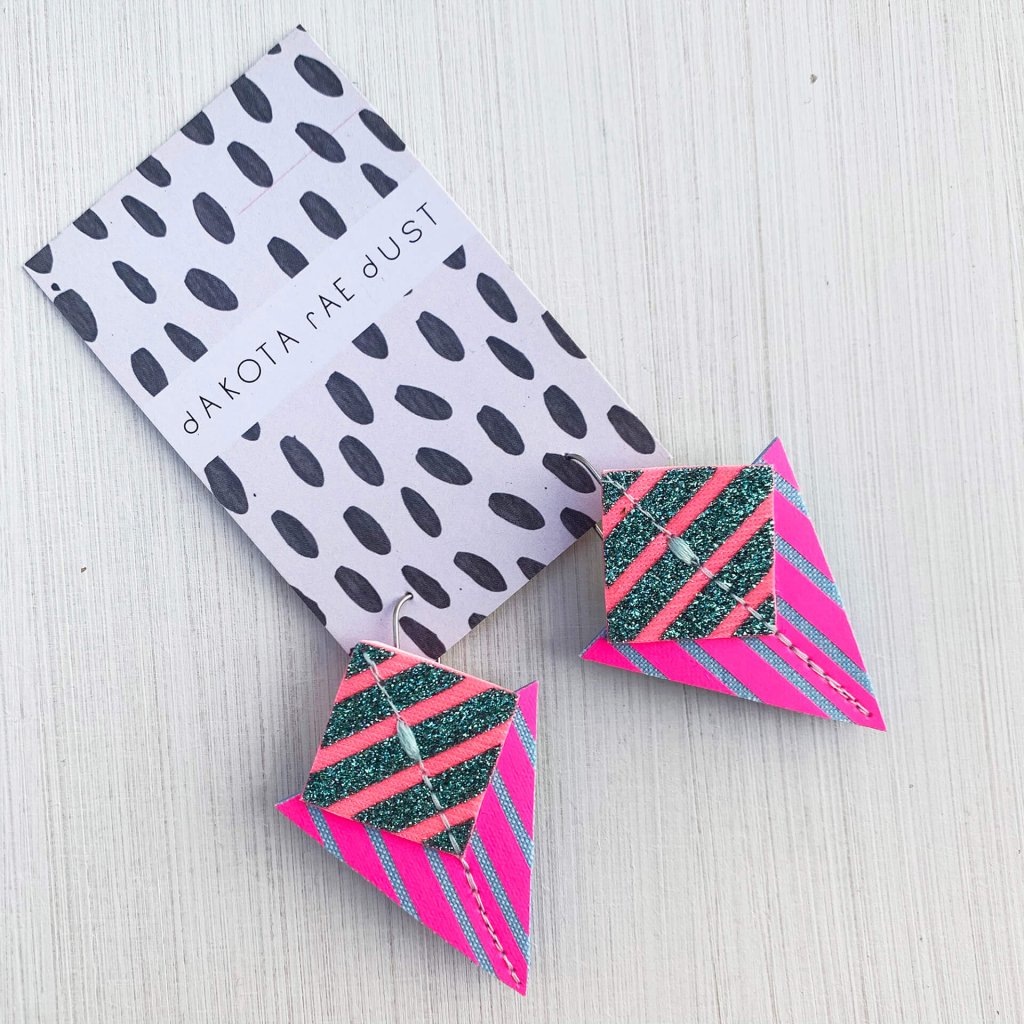 A pair of fluorescent pink, emerald glitter and pale blue Stripey triangle earrings, featuring a triangle and square printed with bold graphic stripes displayed on a black and white patterned, dakota rae dust branded card against an off white background