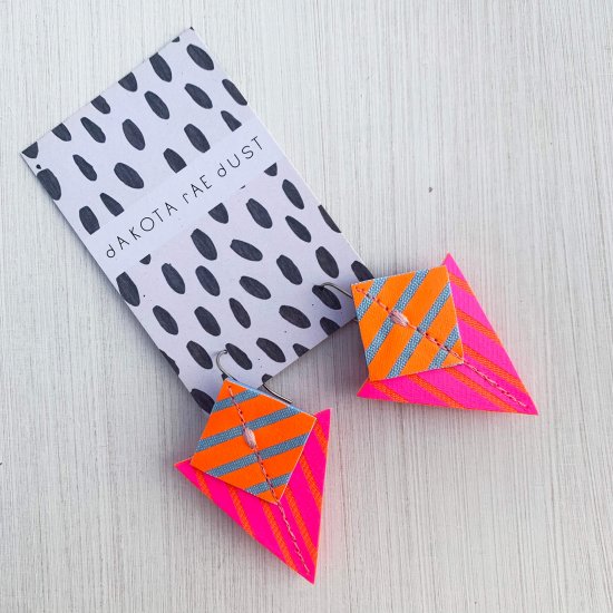 A pair of fluorescent pink, orange and blue Stripey triangle earrings, featuring a triangle and square printed with bold graphic stripes displayed on a black and white patterned, dakota rae dust branded card, against an off white background