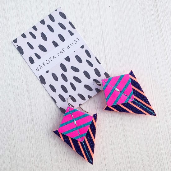 A pair of glittery blue and pink Stripey triangle earrings, featuring a triangle and square printed with bold graphic stripes displayed on a black and white patterned, dakota rae dust branded card, against an off white background