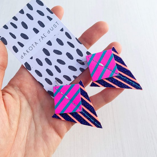 A pair of triangle shaped statement earrings, printed with blue and pink graphic stripes are mounted on a black and white patterned, dakota rae dust branded card and held out for the camera in a white woman's hand