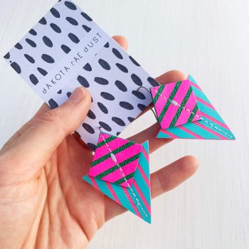 A pair of turquoise, fluorescent pink and green Stripey triangle earrings, featuring a triangle and square printed with bold graphic stripes displayed on a black and white patterned, dakota rae dust branded card, held in a white woman's hand against an off white background