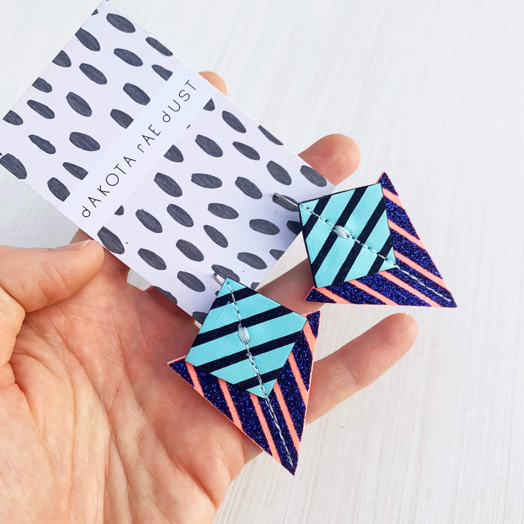 A pair of glittery blue, pale blue, pink and navy Stripey triangle earrings, featuring a triangle and square printed with bold graphic stripes displayed on a black and white patterned, dakota rae dust branded card, held in a white woman's hand against an off white background